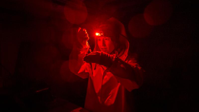 A still from the film Into the Dark. The scene is dark all around. A person in a protective suit and gloves intently examines something in their hands. The only illumination is from a tiny red light mounted on their forehead.
